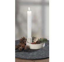 Brandende witte staafkaars in witte houder, versierd met dennenappels, schors en dennentak op grijze vilten onderzetter.