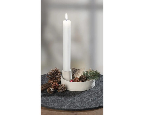 Brandende witte staafkaars in witte houder, versierd met dennenappels, schors en dennentak op grijze vilten onderzetter.