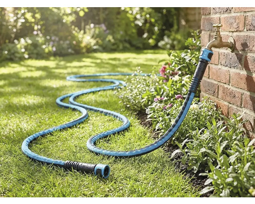 Textiele tuinslang aangesloten op een buitenkraan aan een bakstenen muur, liggend op een gazon in de tuin.
