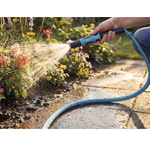 Een hand bewatert bloemen met een for_q tuinsproeikop en een blauwe textielslang in een lichte tuin met stenen tegels.