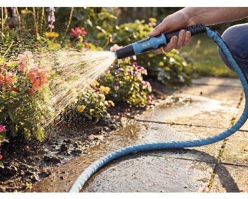 Een hand bewatert bloemen met een for_q tuinsproeikop en een blauwe textielslang in een lichte tuin met stenen tegels.