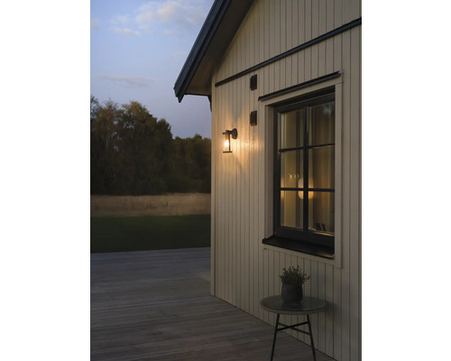Buitenwandlamp op een houten gevel naast een raam bij schemering, houten terras met een bijzettafel.