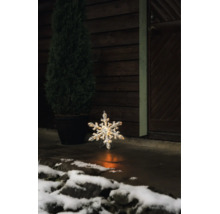 Verlichte LED-sneeuwvlok voor buiten op een stenen vloer voor een donkere houten wand met een plant in pot en sneeuw.