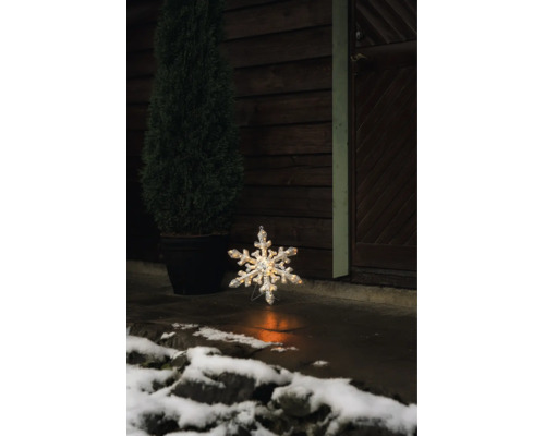 Verlichte LED-sneeuwvlok voor buiten op een stenen vloer voor een donkere houten wand met een plant in pot en sneeuw.