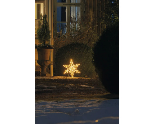 Warmwitte LED-sneeuwvlok als buitenverlichting, geplaatst op de grond in een winterse tuin 's nachts bij een struik en een huis.