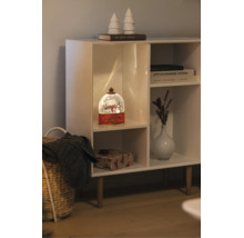 Verlichte sneeuwbol met winters tafereel in een witte kast naast een vaas, boeken en een ingepakt cadeau.