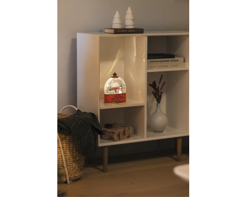 Verlichte sneeuwbol met winters tafereel in een witte kast naast een vaas, boeken en een ingepakt cadeau.