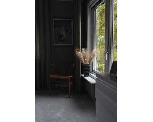 Verlichte pampasgras takken in een vaas op de vensterbank naast een houten stoel in een sfeervolle kamer.