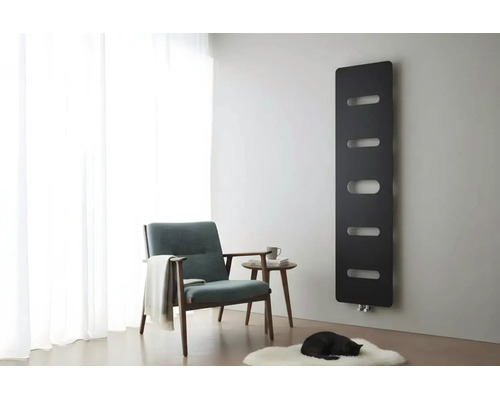 Zwarte designradiator aan een grijze muur in een lichte woonkamer met fauteuil, bijzettafel en wit tapijt.