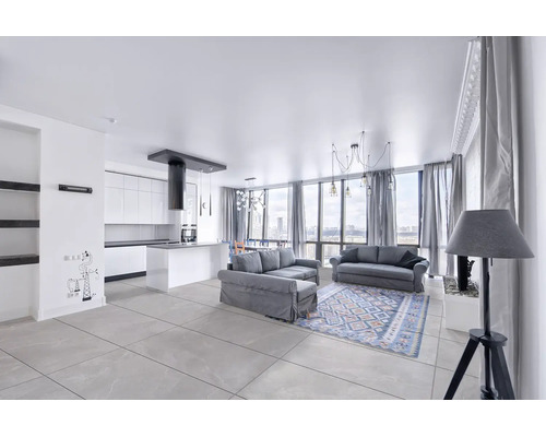 Moderne, lichte open woonkamer en keuken met grijze banken, een gedessineerd tapijt, witte keukenkasten en grijze vloertegels.