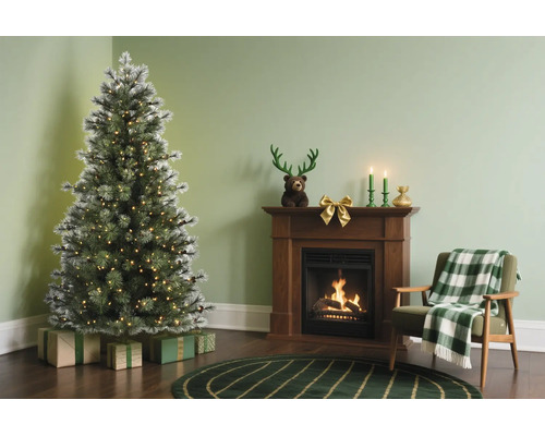 Kerstachtig ingerichte woonkamer met een kunstmatige besneeuwde kerstboom met lichtjes naast een houten haard en een fauteuil.