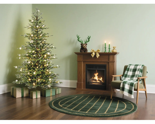 Feestelijke woonkamer met een verlichte kerstboom, houten haard, groen vloerkleed en fauteuil met geruite deken.