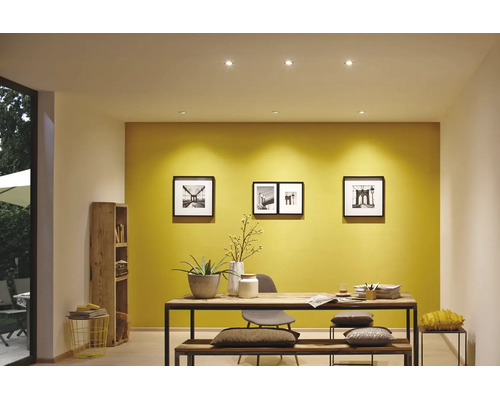 Een modern ingerichte kamer met gele muurverf, ronde plafondinbouwspots en een massief houten tafel met bankje.
