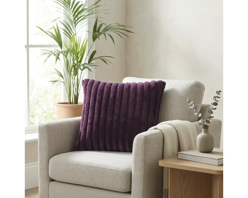 Paars geribbeld sierkussen op een lichtgrijze fauteuil in een lichte kamer met kamerplant en bijzettafel.