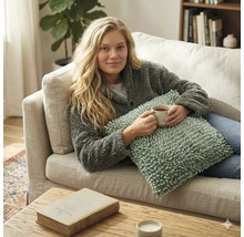 Vrouw op een bank in een lichte woonkamer met een getextureerd sierkussen en mok in haar handen, gezellige sfeer met plant en boekenkast.