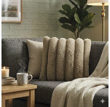 Beige sierkussen van imitatiebont met verticale ribbels op een grijze bank in een woonkamer met gebreide deken en mok op een bijzettafel.