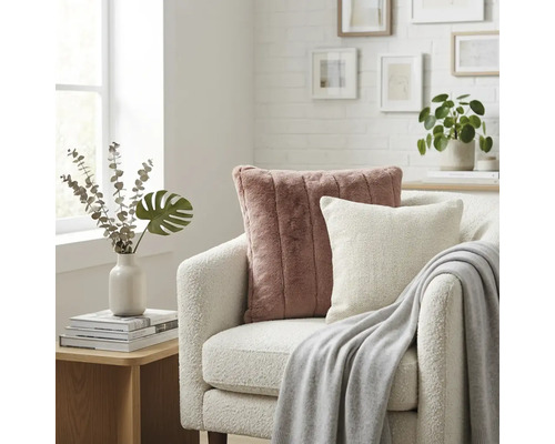 Lichte woonkamer met een witte bouclé fauteuil, een oudroze imitatiebont kussen, een wit sierkussen en een grijze plaid.