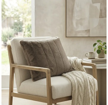 Vierkant, geribbeld sierkussen van imitatiebont op een lichte fauteuil met een gebreide deken in een lichte, modern ingerichte kamer.