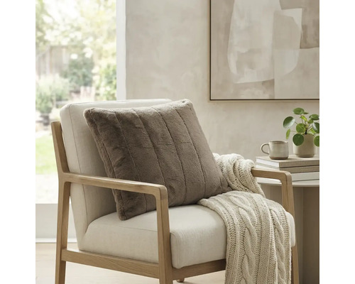 Vierkant, geribbeld sierkussen van imitatiebont op een lichte fauteuil met een gebreide deken in een lichte, modern ingerichte kamer.