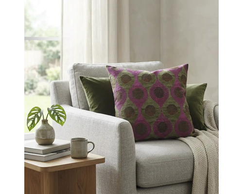 Sierkussen met paars en groen geometrisch patroon op een lichtgrijze fauteuil in een lichte woonkamer met kamerplant.