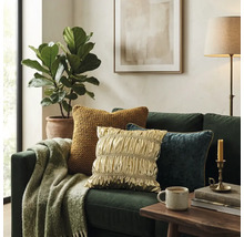 Groene bank met kussens in goud, blauw en geel, een deken met patroon, houten tafel met mok, plant en vloerlamp in een lichte kamer.