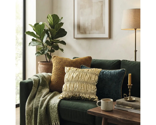 Groene bank met kussens in goud, blauw en geel, een deken met patroon, houten tafel met mok, plant en vloerlamp in een lichte kamer.