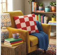 Sierkussen met ruitpatroon op een beklede fauteuil met een gebreide plaid in een lichte woonkamer met boekenkast en lamp.