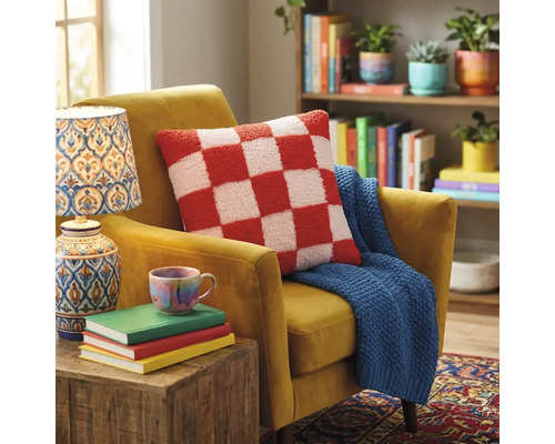 Sierkussen met ruitpatroon op een beklede fauteuil met een gebreide plaid in een lichte woonkamer met boekenkast en lamp.