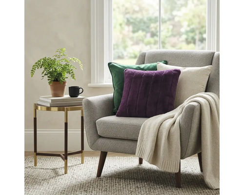 Licht ingerichte woonkamer met fauteuil, sierkussens en plaid. Daarnaast een ronde bijzettafel met plant en mok op een tapijt.