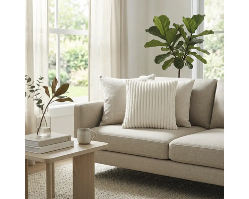 Lichte woonkamer met beige stoffen bank, wit ribfluwelen sierkussen en houten bijzettafel. Grote kamerplant en raam op de achtergrond.