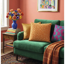 Groene fluwelen bank met luipaardprint kussen, een oranje deken, een bijzettafel met bloemen en ingelijste wandkunst.