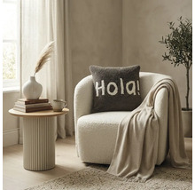 Een licht ingerichte woonkamer met een witte bouclé fauteuil, een bruin sierkussen met de tekst Hola en een geribbelde bijzettafel.