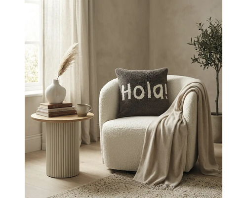 Een licht ingerichte woonkamer met een witte bouclé fauteuil, een bruin sierkussen met de tekst Hola en een geribbelde bijzettafel.