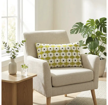 Een lichte woonkamer met een beige gestoffeerde fauteuil met een groen sierkussen met wit bloemenpatroon, naast een Monstera plant.