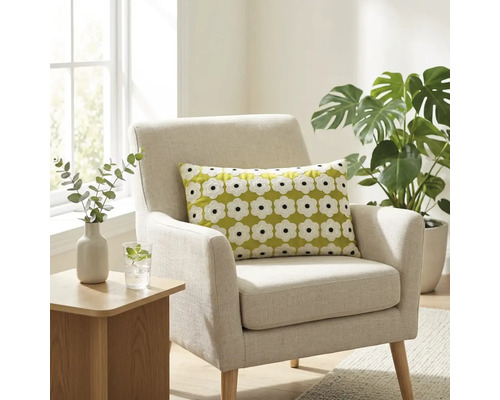 Een lichte woonkamer met een beige gestoffeerde fauteuil met een groen sierkussen met wit bloemenpatroon, naast een Monstera plant.