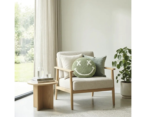 Lichte woonkamer met houten fauteuil, rond groen smiley-kussen, bijzettafel en kamerplant voor een raam met gordijnen.