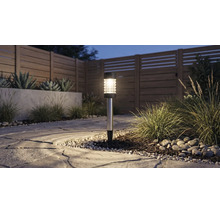Solarlamp in de tuin op een stenen pad naast siergrassen en een houten schutting in de schemering.