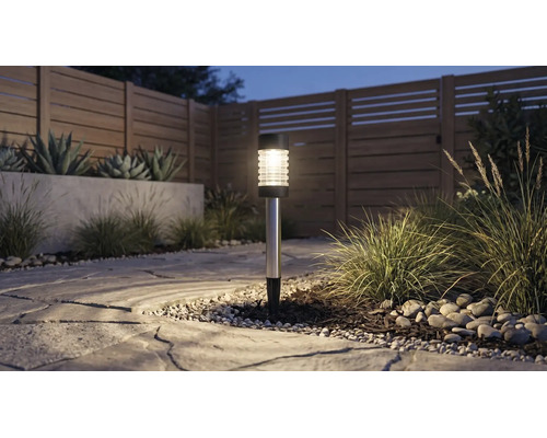 Solarlamp in de tuin op een stenen pad naast siergrassen en een houten schutting in de schemering.