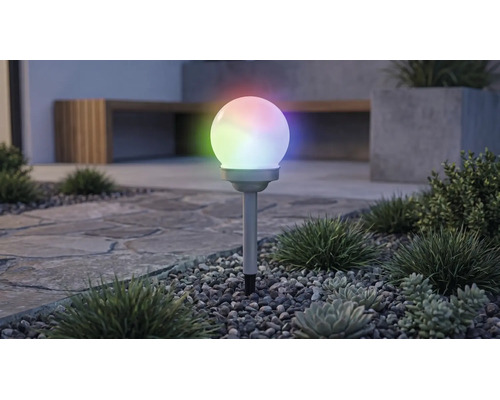 Tuinverlichting op zonne-energie in bolvorm met meerkleurige RGB-verlichting, op een grondpen in een grindbed met planten.