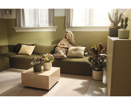Lichte woonkamer met olijfgroene bank, vrouw met mok, beige salontafel en diverse kamerplanten in een natuurlijke stijl.