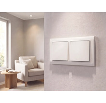 Moderne witte dubbele lichtschakelaar op een getextureerde wand in een lichte woonkamer met een fauteuil en bijzettafel.