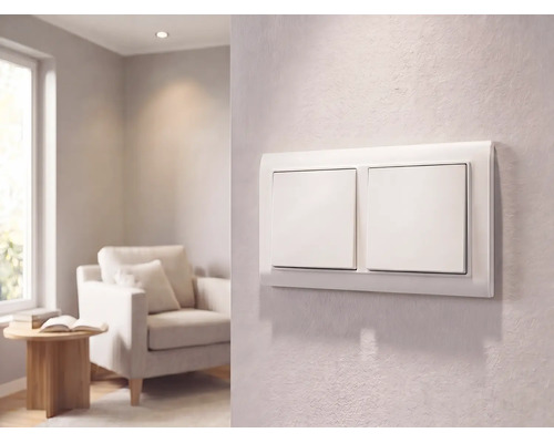 Moderne witte dubbele lichtschakelaar op een getextureerde wand in een lichte woonkamer met een fauteuil en bijzettafel.