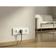 Drievoudig stopcontact met een ingestoken stekker aan een muur boven een houten vloer in een lichte woonkamer.