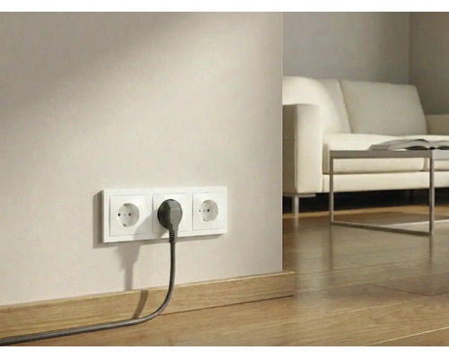 Drievoudig stopcontact met een ingestoken stekker aan een muur boven een houten vloer in een lichte woonkamer.