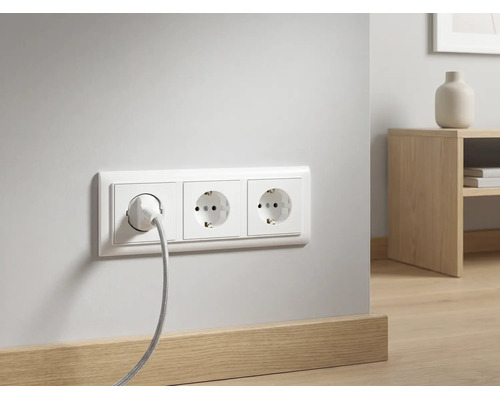 Drievoudig stopcontact met een aangesloten kabel op een witte muur boven een houten plint in een moderne kamer.