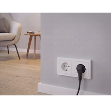 Dubbel stopcontact met aangesloten stroomkabel, op de achtergrond een lichte woonkamer met fauteuil, bijzettafel en houten vloer.