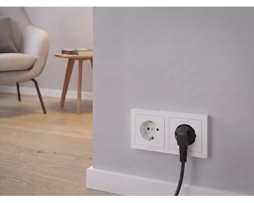 Dubbel stopcontact met aangesloten stroomkabel, op de achtergrond een lichte woonkamer met fauteuil, bijzettafel en houten vloer.