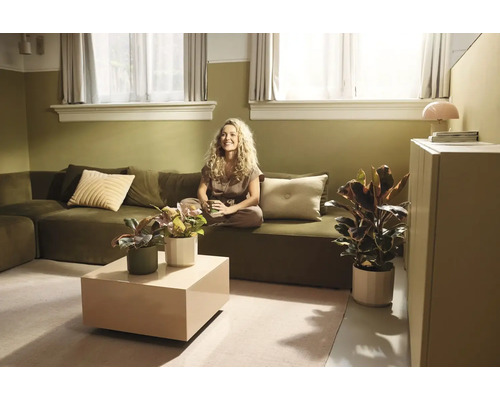 Lichte woonkamer met een olijfgroene hoekbank, een beige salontafel en verschillende kamerplanten in potten.