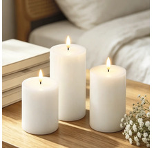 Drie witte LED-stompkaarsen van verschillende hoogtes op een houten tafel naast boeken en bloemen.