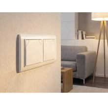 Dubbele lichtschakelaar op een muur in een lichte woonkamer met bank en vloerlamp.